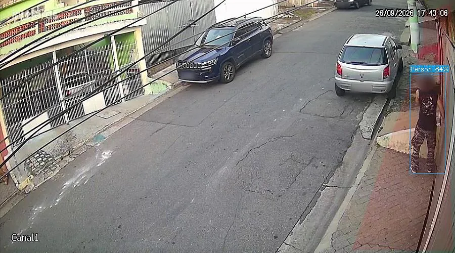 Imagem real de câmera em uma rua residencial: a inteligência artificial marca a pessoa parada junto ao portão da casa. Rosto e placas desfocados.